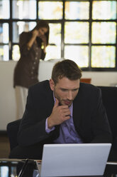 Man in office using laptop, woman in background phoning - WESTF10676