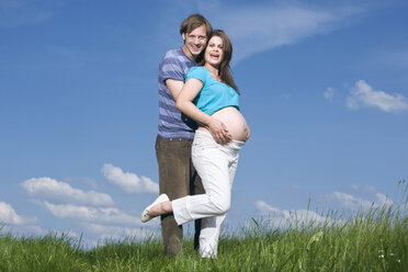 Junger Mann, der eine schwangere Frau umarmt, auf einer Wiese stehend, lächelnd, Porträt - RDF00963