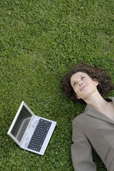 Junge Geschäftsfrau auf einer Wiese liegend mit einem Laptop neben sich, Blick von oben, Porträt - KJF00063