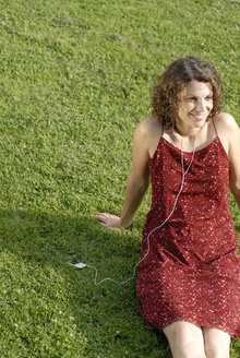 Young woman sitting on lawn, listening to MP3 player, smiling - KJF00057