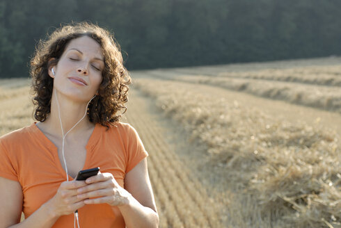 Young woman in field listening to MP3 player, portrait - KJF00050