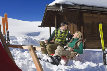 Italy, South Tyrol, Seiseralm, Couple sitting in front of log cabin, holding champagne glasses, smiling, portrait - WESTF11438