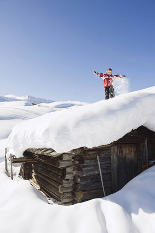 Italien, Südtirol, Seiseralm, Junge (4-5) auf schneebedecktem Dach einer Blockhütte stehend, lizenzfreies Stockfoto