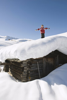 Italien, Südtirol, Seiseralm, Junge (4-5) auf schneebedecktem Dach einer Blockhütte stehend - WESTF11398