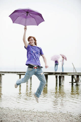 Deutschland, Bayern, Ammersee, Junger Mann hält Regenschirm und springt, lizenzfreies Stockfoto