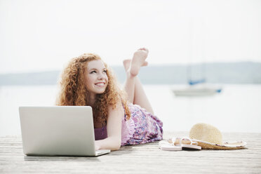 Germany, Bavaria, Ammersee, Young woman lying on jetty using laptop, portrait - WESTF12220