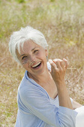 Spain, Mallorca, Senior woman sitting in meadow, holding blade of grass, laughing, elevated view - WESTF12941