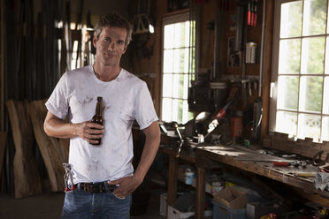 Germany, Hamburg, Man in home workshop holding beer bottle - WEST13141