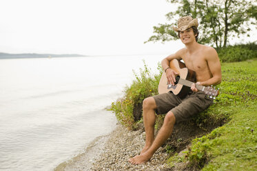 Deutschland, Bayern, Starnberger See, Junger Mann sitzt am Seeufer und spielt Gitarre, lächelnd, Porträt - RNF00047