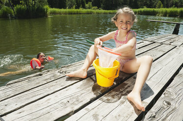 Italy, South Tyrol, Girl (6-7) playing with net and bucket on jetty, girl (8-9) in background swimming in lake - WESTF13359