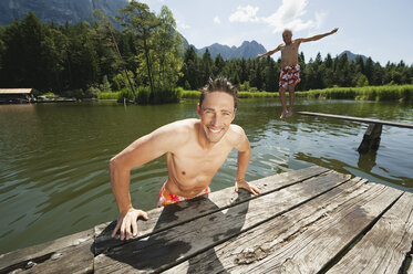 Italy, South Tyrol, Man in foreground leaning on jetty, senior man in background jumping into lake, portrait - WESTF13351