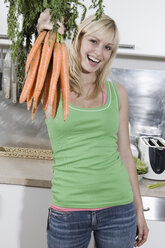 Germany, Berlin, Young woman in kitchen holding bunch of carrots, portrait - WESTF13469