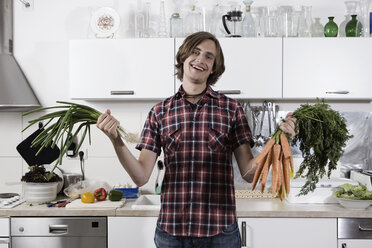 Deutschland, Berlin, Junger Mann in Küche mit Karotten und Frühlingszwiebeln, lachend, Porträt - WESTF13464