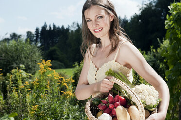 Deutschland, Bayern, Junge Frau hält Korb mit Gemüse im Garten, lächelnd, Porträt - WESTF13241