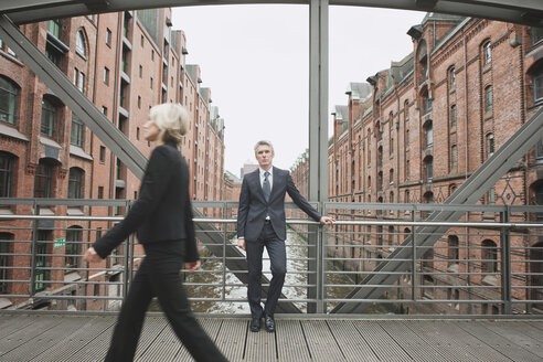 Deutschland, Hamburg, Geschäftsfrau beim Überqueren einer Brücke, Geschäftsmann im Hintergrund lehnt sich an ein Geländer - WESTF13865
