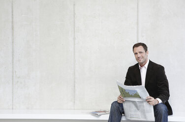 Germany, Cologne, Businessman sitting on bench holding newspaper, smiling, portrait - JOF00061