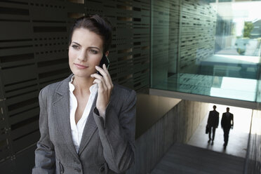 Germany, Cologne, Businesswoman on staircase using mobile phone, businessmen in background - JOF00045