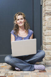 Germany, Cologne, Woman sitting in front of door using laptop, smiling, portrait - WESTF14347