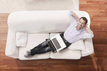 Deutschland, Köln, Mann mit Laptop entspannt auf Sofa, Blick von oben - WESTF14305