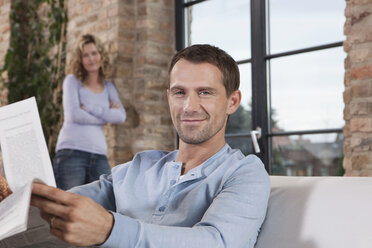 Germany, Cologne, Couple, man on sofa holding newspaper, woman standing in background, portrait, close-up - WESTF14238