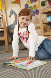 Deutschland, Mädchen (6-7) im Kinderzimmer mit Buch schaut in die Kamera, lächelnd, Porträt - RNF00100