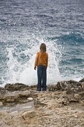 Kroatien, Korcula, Mädchen (8-9) steht am Meer und schaut auf eine plätschernde Welle, Rückansicht - GNF01183