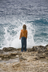Kroatien, Korcula, Mädchen (8-9) steht am Meer und schaut auf eine plätschernde Welle, Rückansicht - GNF01182