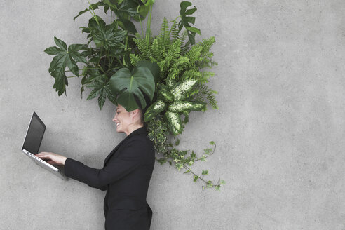 Businesswoman using laptop, foliage plants on head, smiling, portrait, elevated view - BAEF00069