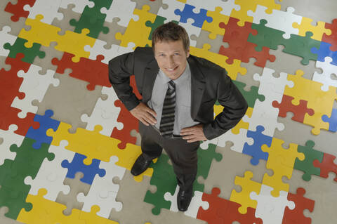 Geschäftsmann auf Puzzle stehend, Hände in die Hüften gestemmt, lächelnd, Porträt, Blick von oben, lizenzfreies Stockfoto