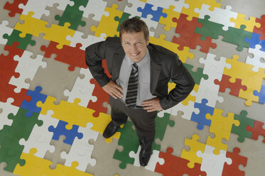 Geschäftsmann auf Puzzle stehend, Hände in die Hüften gestemmt, lächelnd, Porträt, Blick von oben - ASF03965