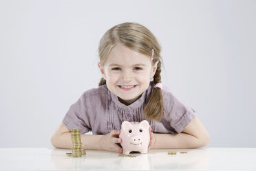 Mädchen (4-5) hält Sparschwein und Stapel Münzen, lächelnd, Porträt - RBF00234