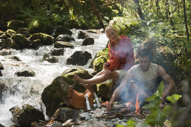 Österreich, Steiermark, Junges Paar sitzt am Lagerfeuer am Bach im Wald - HHF03267
