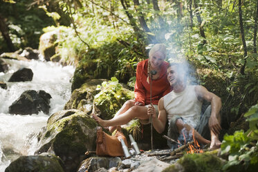 Austria, Steiermark, Young couple sitting at camp fire by stream in forest - HHF03266