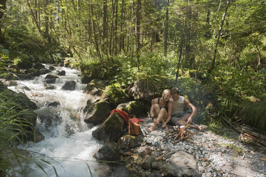 Austria, Steiermark, Young couple sitting at camp fire by stream in forest - HHF03265
