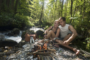 Austria, Steiermark, Young couple sitting at camp fire by stream in forest - HHF03264