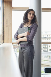 Germany, Leipzig, Young woman standing with books, looking away - BABF00573