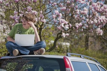 Deutschland, Junge sitzt auf dem Autodach mit Laptop und Magnolienbaum im Hintergrund - WDF00737