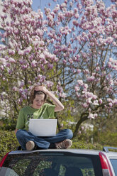 Germany, Boy sitting on car roof and using laptop with magnolia tree in background - WDF00736