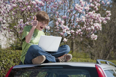 Deutschland, Junge sitzt auf dem Autodach und benutzt Laptop mit Magnolienbaum im Hintergrund - WDF00735