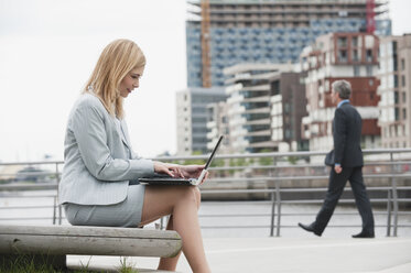 Germany, Hamburg, Businesswoman using laptop, man walking in background - WESTF015416