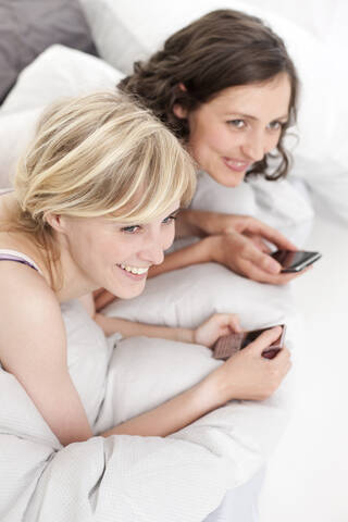 Deutschland, Leipzig, Junge Frau auf Bett mit Handy, lächelnd, lizenzfreies Stockfoto