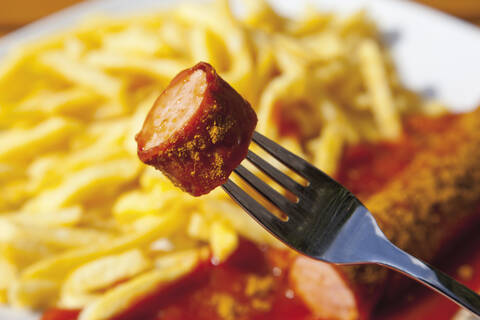 Currywurst auf Gabel mit Pommes im Hintergrund, lizenzfreies Stockfoto