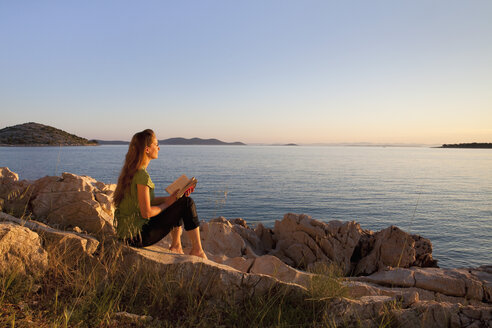 Kroatien, Zadar, Junge Frau mit Buch und Blick auf den Strand - HSIF000099