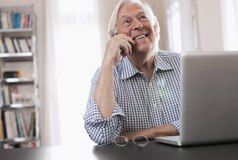 Deutschland, Wakendorf, Älterer Mann am Telefon mit Laptop, lächelnd, lizenzfreies Stockfoto