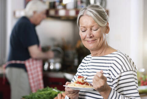 Deutschland, Wakendorf, Ältere Frau isst Nudeln, Mann kocht im Hintergrund, lizenzfreies Stockfoto