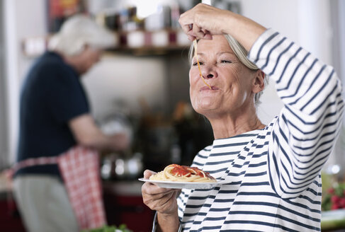 Germany, Wakendorf, Senior woman eating noodles, man cooking in background - WESTF016255