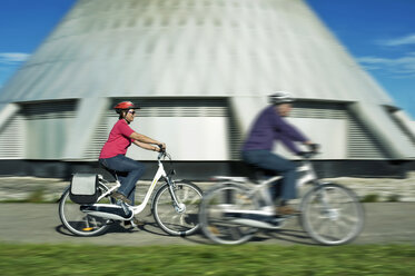 Deutschland, Bayern, Raisting, Älteres Paar fährt Elektrofahrrad in der Nähe eines Radiosenders - RNF000659