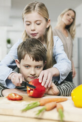 Germany, Cologne, Girl teaching boy cutting vegetables, mother in background - WESTF016347