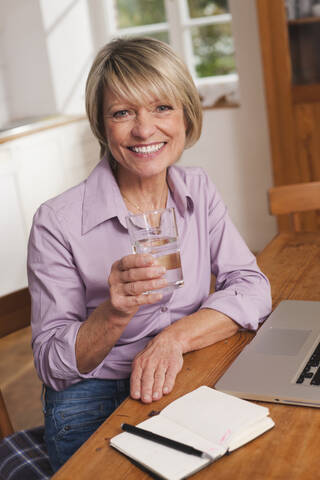 Deutschland, Kratzeburg, Seniorin mit Wasserglas und Laptop, lächelnd, Porträt, lizenzfreies Stockfoto
