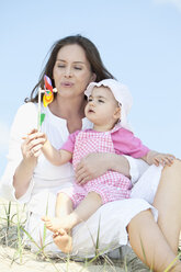 Germany, Bavaria, Mother blowing paper windmill with baby girl sitting on her lap - MAEF003249